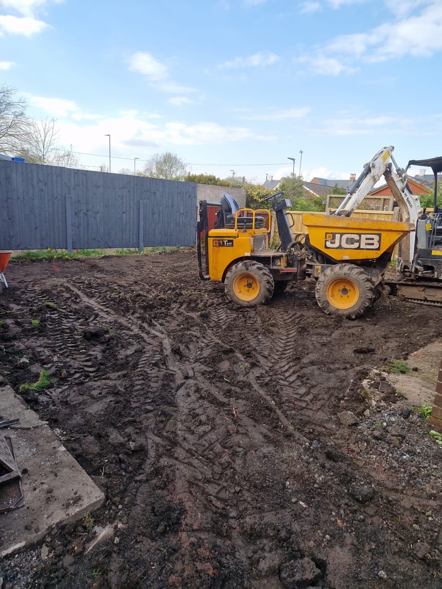 LUNA SEN-Scapes garden dig off: yellow high-tip site dumper and mini excavator on excavated wet soil in a fenced UK family garden, ready for formation level