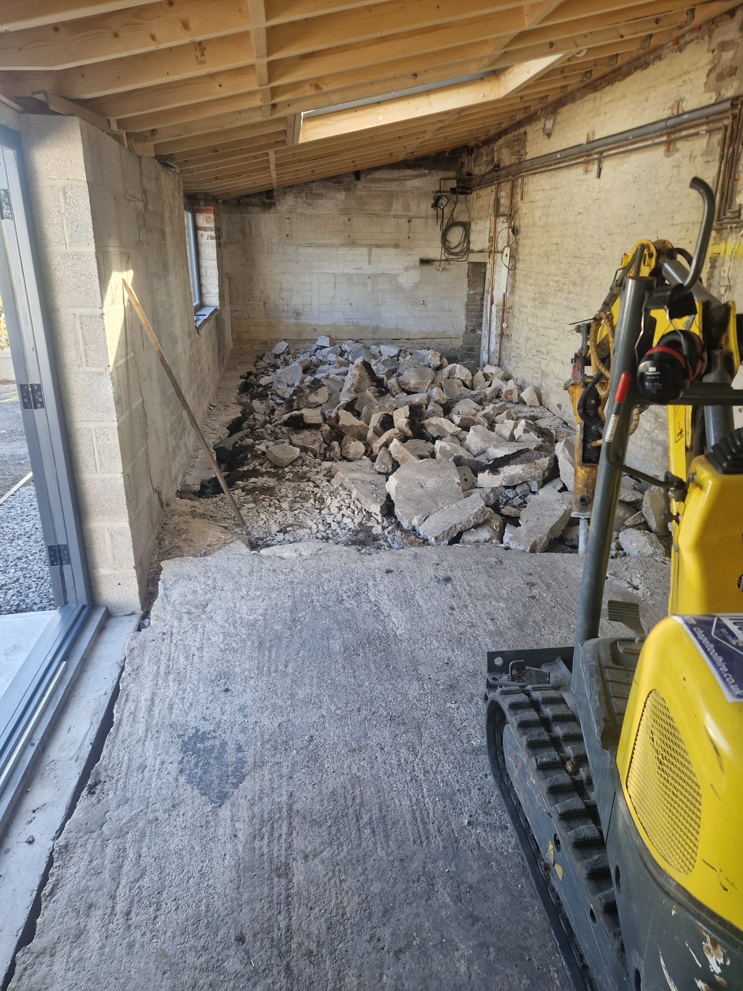 Internal concrete floor dig out by LUNA SEN-Scapes: yellow mini excavator, broken slab stacked on the sub-base, new blockwork and bi-fold opening under exposed timber rafters