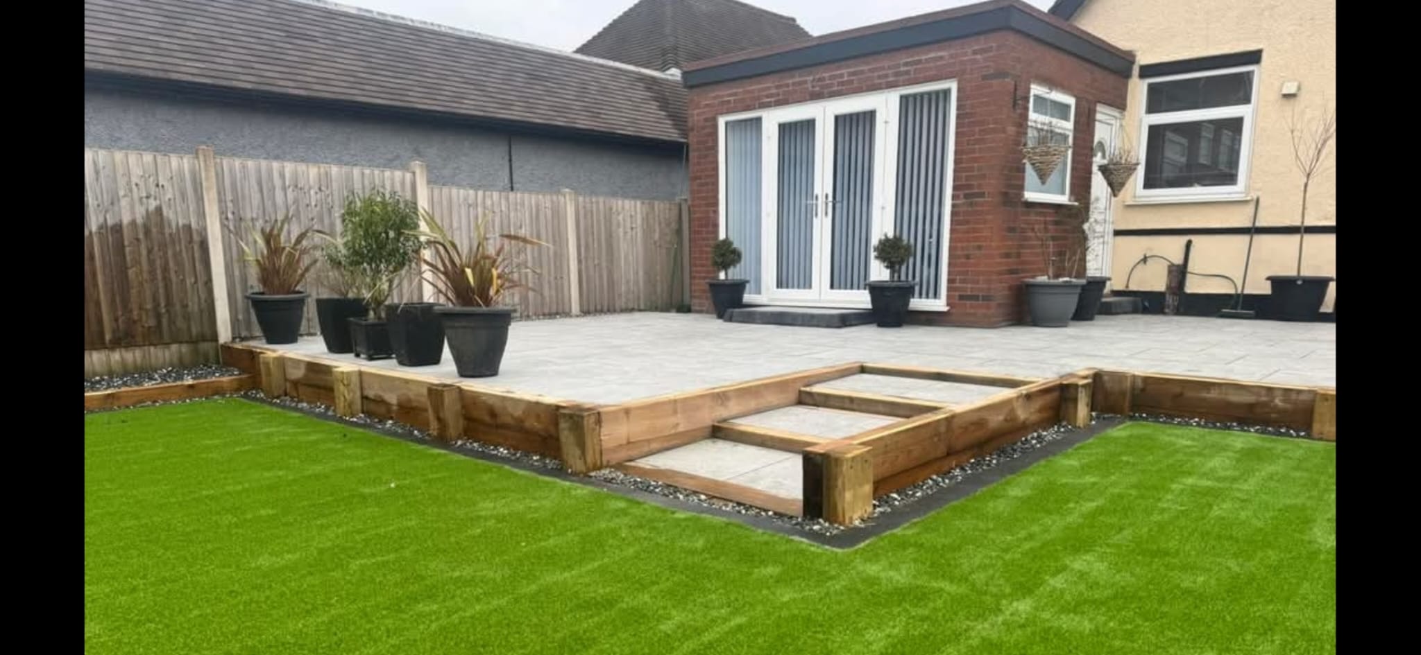 Finished accessible garden makeover by LUNA SEN-Scapes: raised light-grey patio, timber sleeper retainers, shallow paved steps and a level artificial lawn