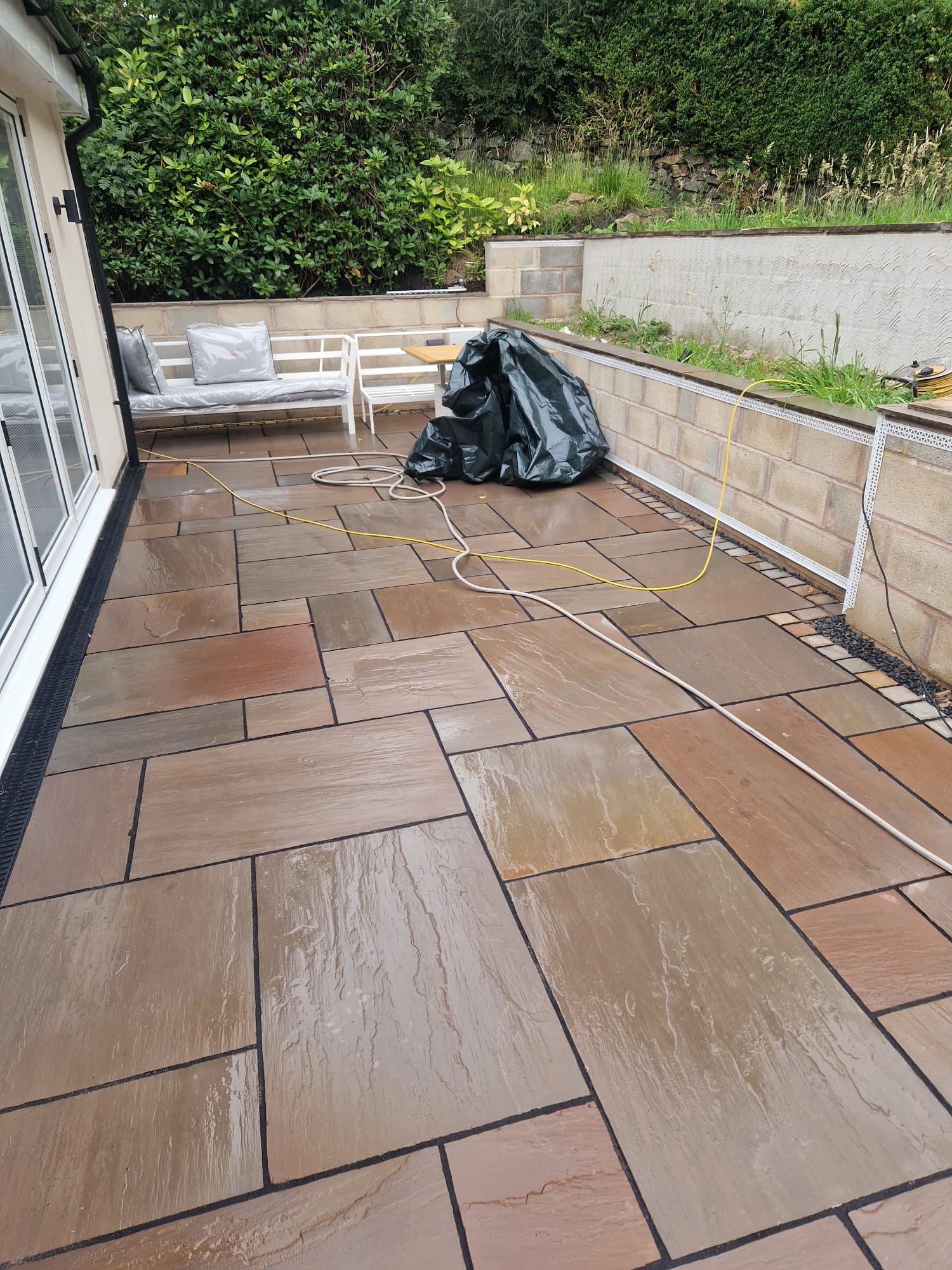 Newly laid multi-tonal Indian sandstone patio flush with white sliding doors, with a black ACO drainage channel and cobble sett edging by LUNA SEN-Scapes