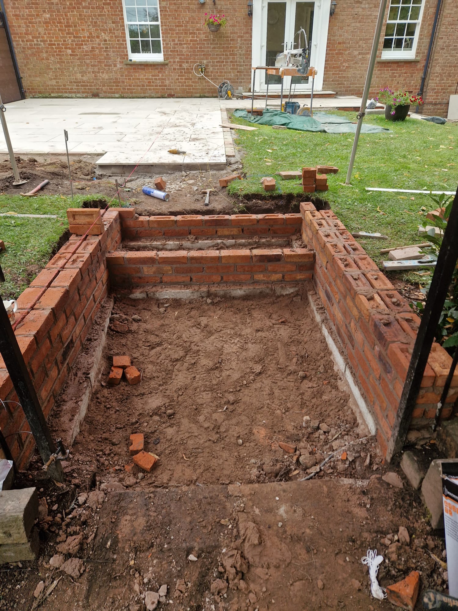 Garden groundworks in progress by LUNA SEN-Scapes: rectangular excavation lined with red brick step walls, string lines for level, and a light stone patio forming beside a brick house